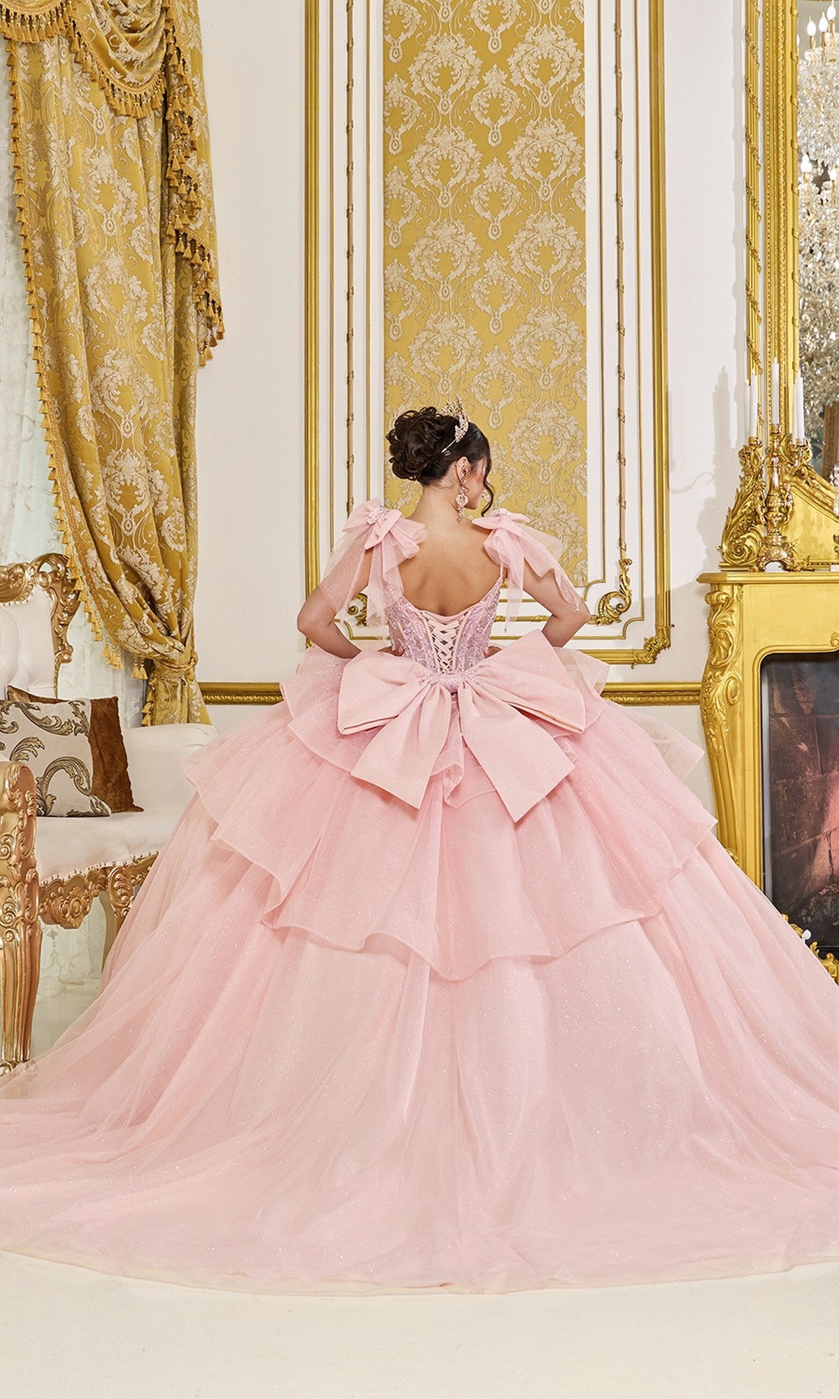 A woman wears the Quinceanera Dress 5079 By Dancing Queen, featuring blush pink fabric and a large bow, as she stands with her back to the camera in an ornate room with gold accents.